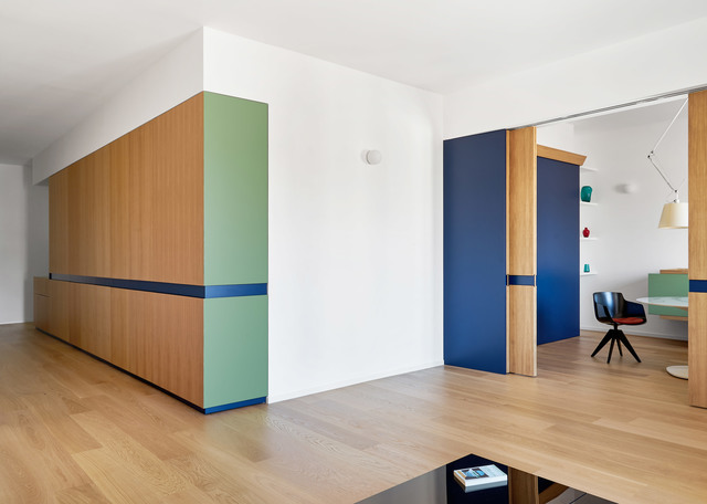 Interno moderno con mobili in legno dai colori blu e verde, apertura su uno spazio lavoro con lampada a sospensione e lampada da tavolo.