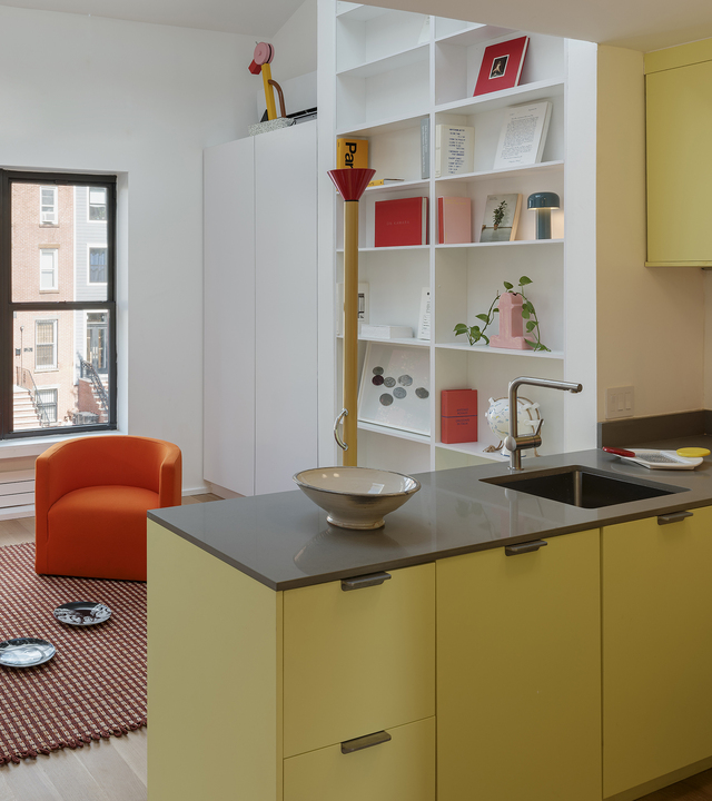 Cucina moderna con pensili gialli e piano grigio, libreria aperta con libri colorati e una lampada da tavolo rosa