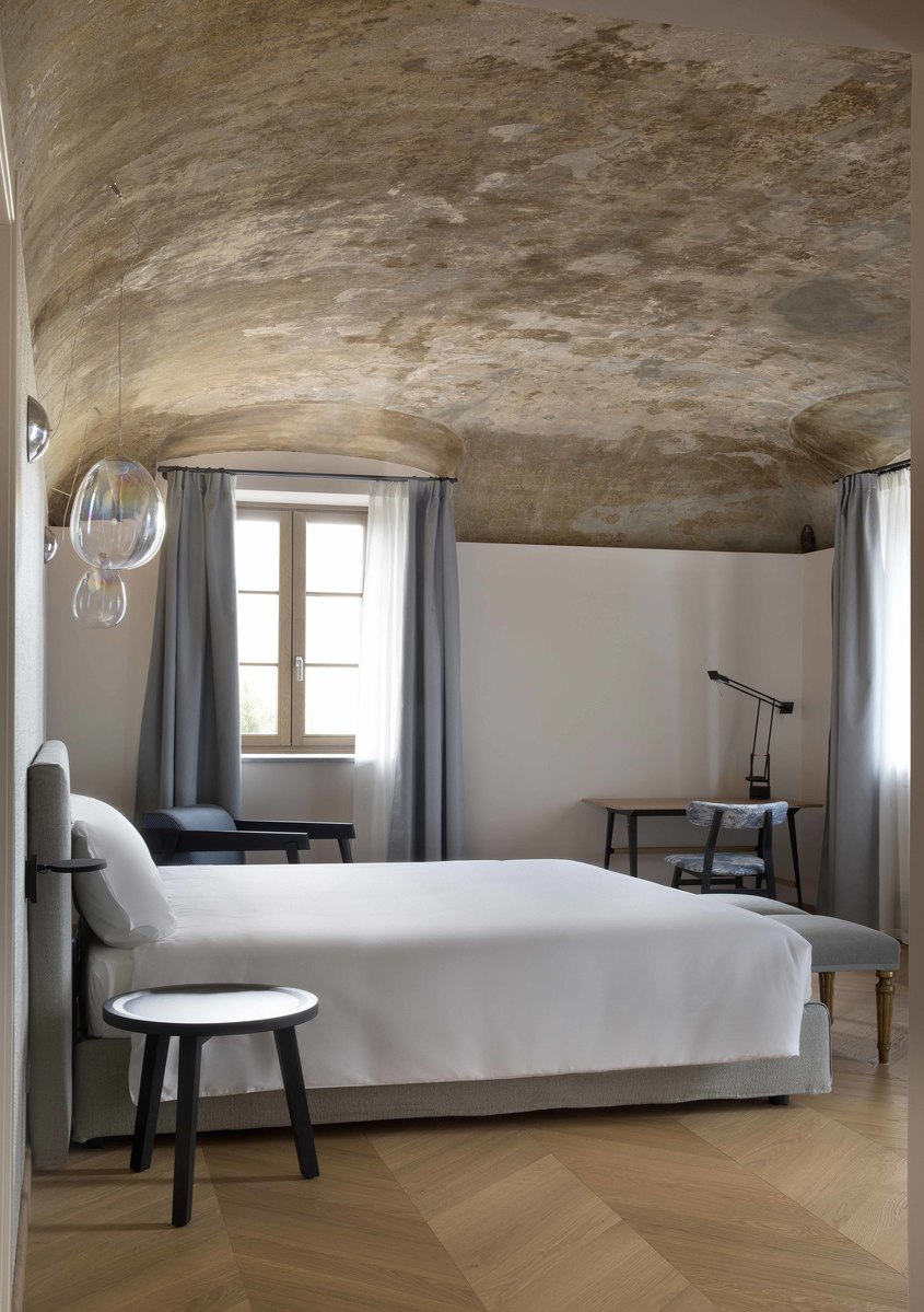 Elegant bedroom with vaulted stone ceiling, white bed, herringbone floor, Artemide Tolomeo desk lamp, and a spherical glass pendant light