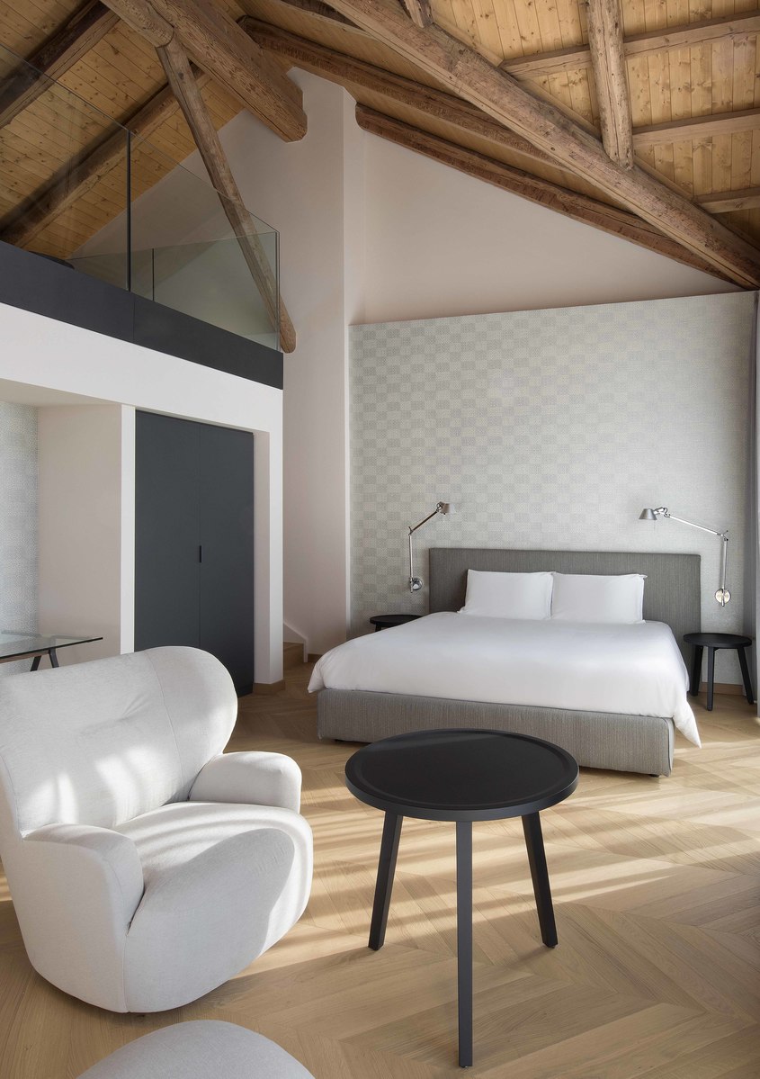 Hotel bedroom with wooden beamed ceiling, white armchair, black coffee table, and two Artemide bedside lamps on nightstands flanking a gray upholstered bed.