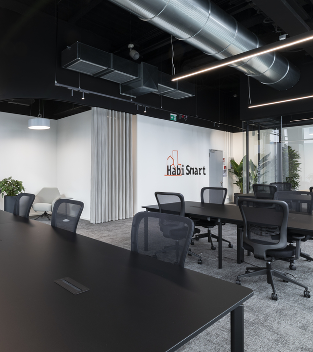 Ufficio moderno con scrivanie nere, sedie a rete, soffitto con condotti a vista, lampade a sospensione LED lineari e logo HabiSmart su parete bianca.