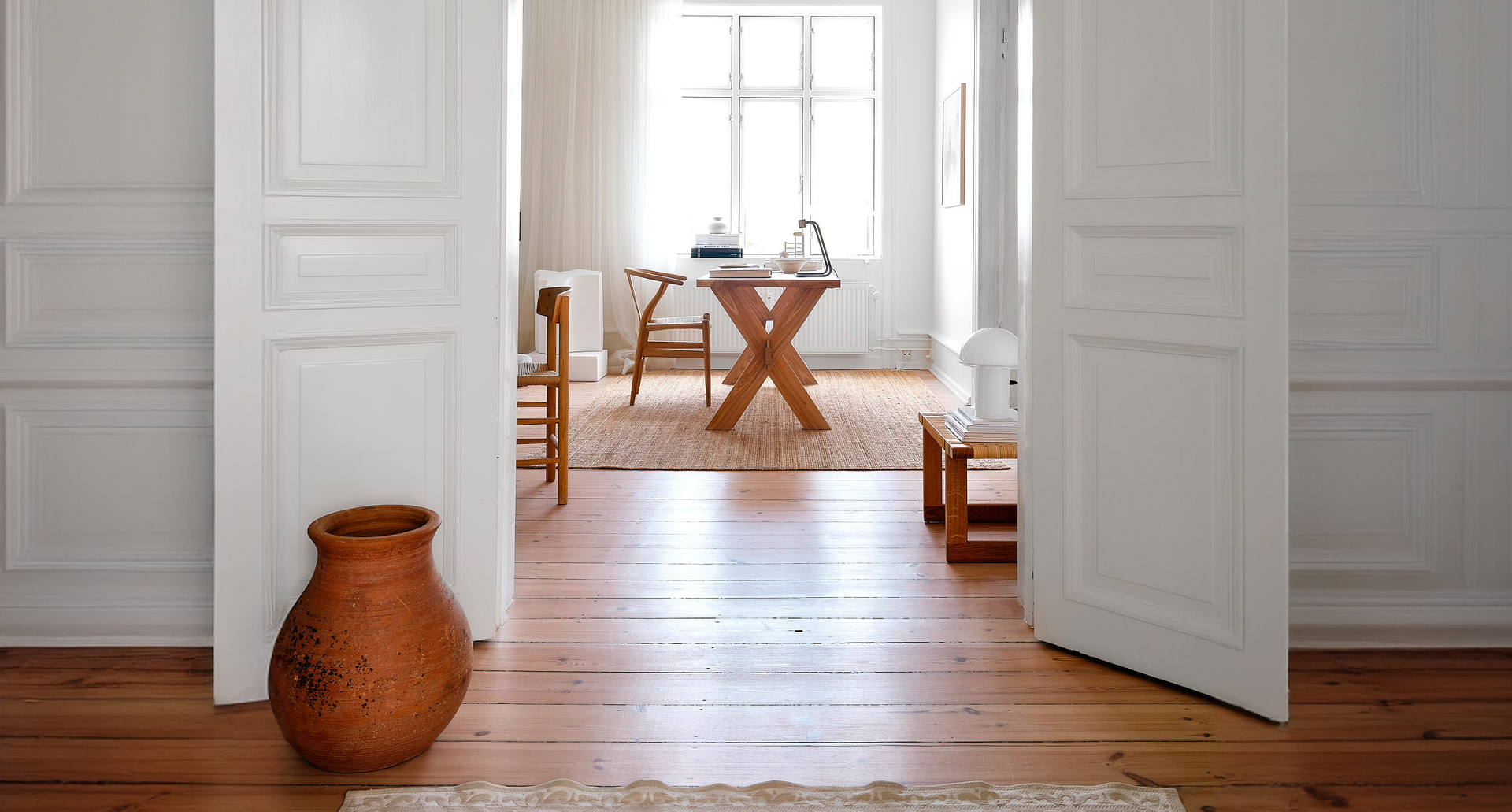 Bright Scandinavian-style room with wooden floor, X-leg dining table, wicker chairs, and a small white table lamp by the window