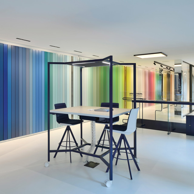 Spazio di lavoro moderno con tavolo alto, sgabelli sotto una struttura nera a baldacchino, luci a soffitto incassate accanto a una parete a strisce multicolori.