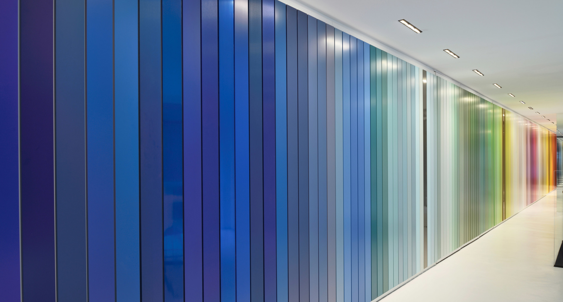 Corridoio con parete a gradiente cromatico dal pavimento al soffitto, che va dal blu intenso al verde e toni caldi, illuminata da faretti a incasso.