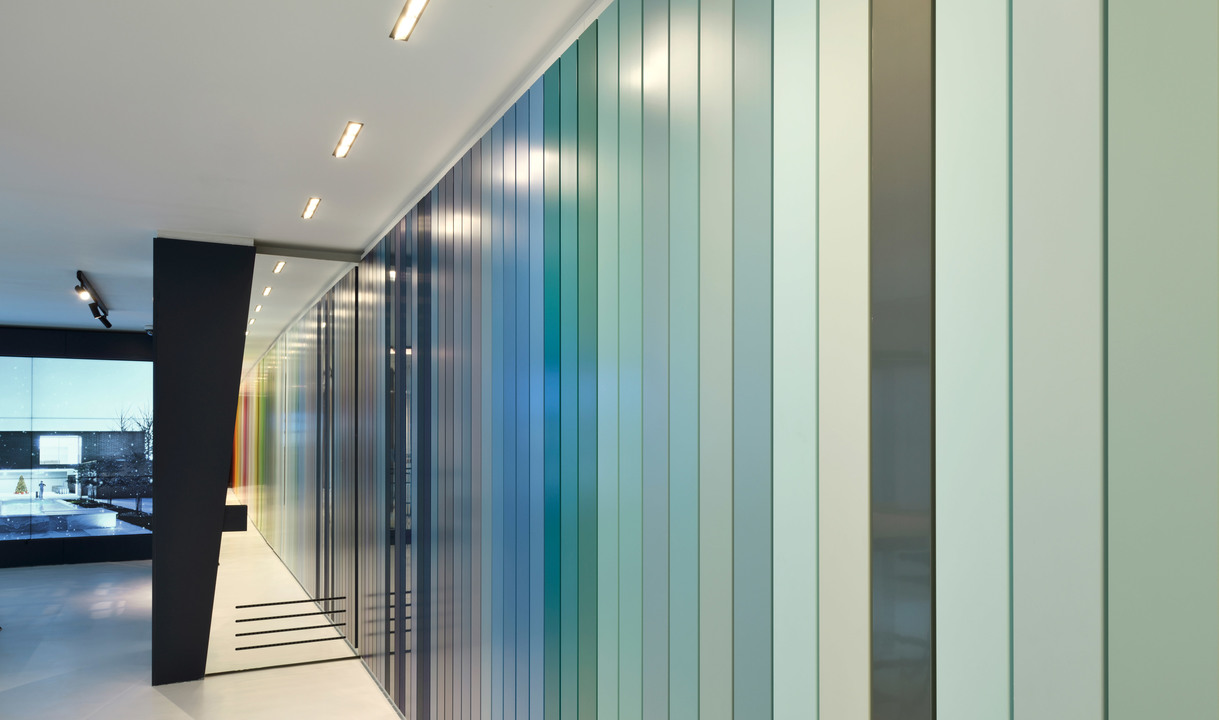 Corridoio moderno con grande parete illuminata di pannelli in vetro verticali multicolori, che sfumano dal blu al verde, illuminati da faretti a soffitto incassati.