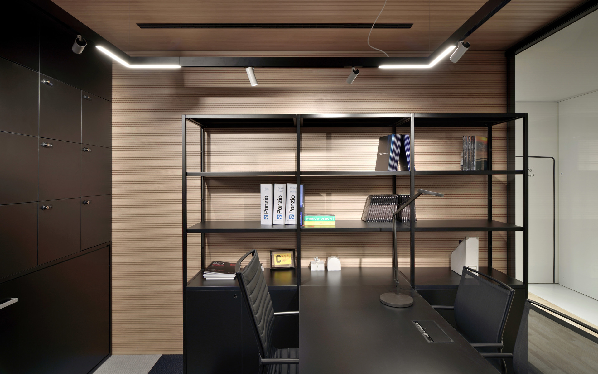 Ufficio moderno con scrivania scura, libreria aperta e due sottili lampade lineari a LED a soffitto che illuminano lo spazio di lavoro.