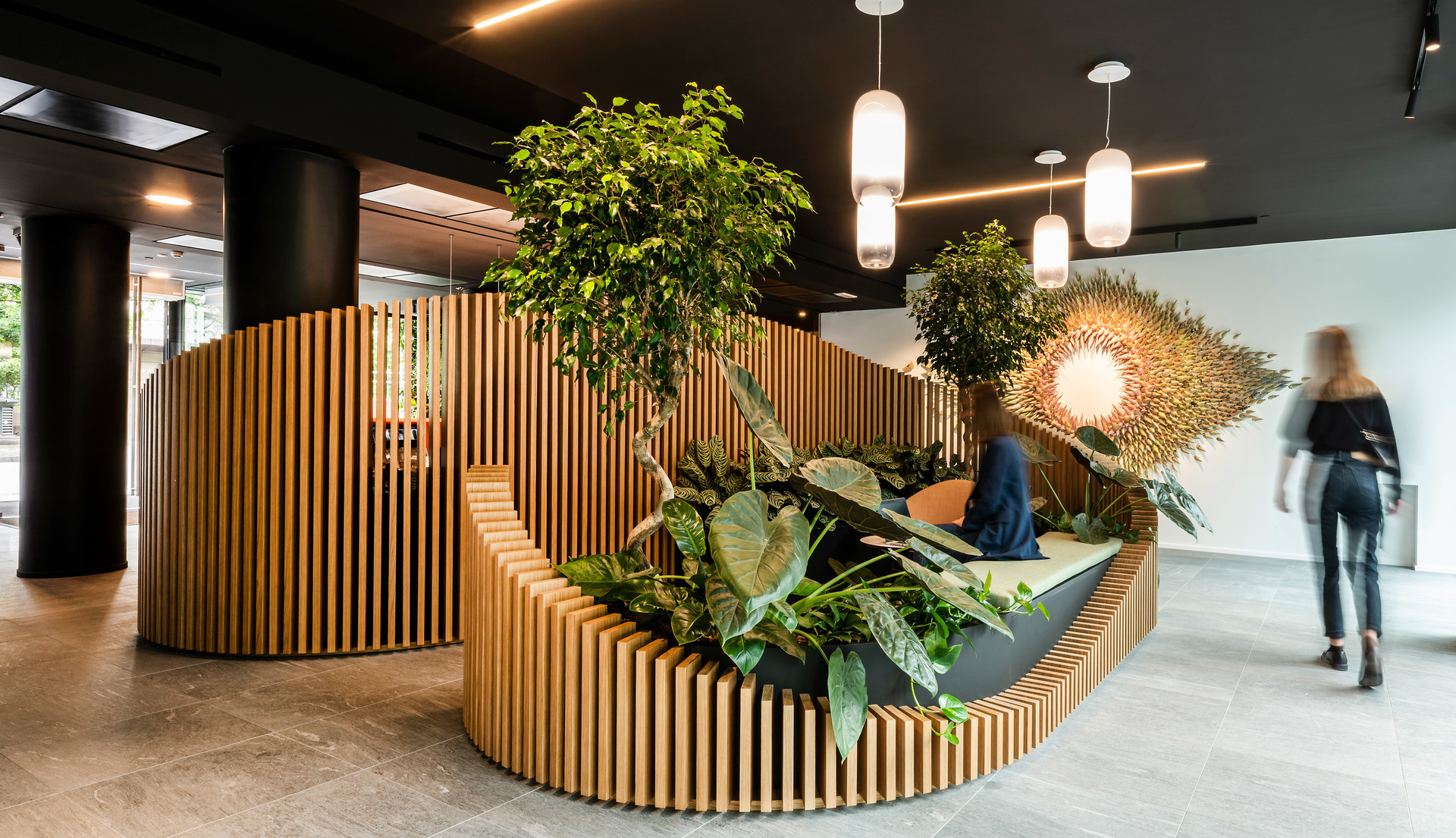 Ingresso ufficio moderno con sedute in listelli di legno curvi, rigogliosa vegetazione e lampade a sospensione cilindriche Artemide su soffitto scuro
