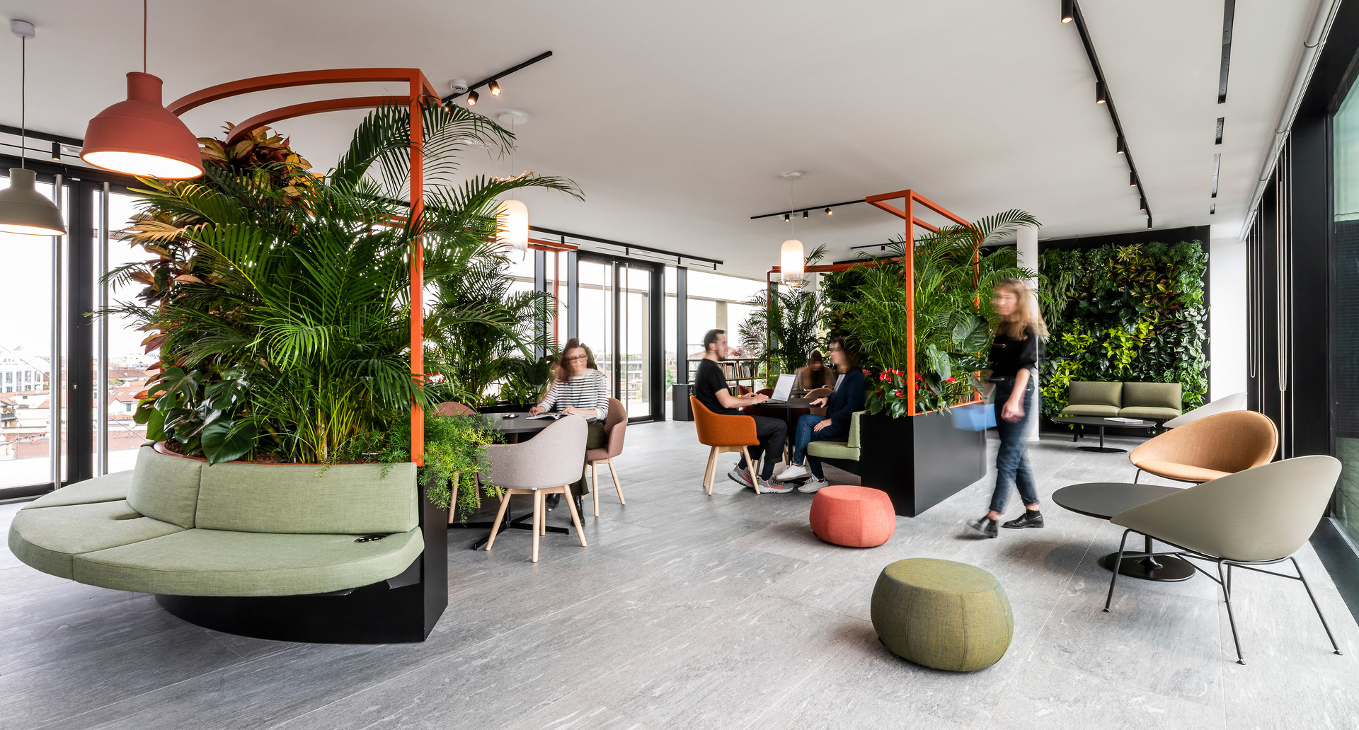 Lounge ufficio moderno con design biofilico, rigogliosa vegetazione, strutture arancioni, illuminazione su binario e aree collaborative dove le persone lavorano e socializzano