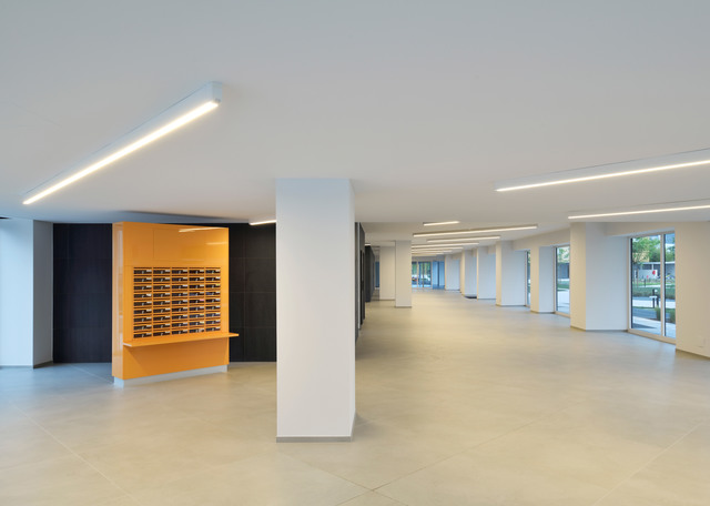 Spacious modern interior with recessed linear LED ceiling lights illuminating an open-plan space with white columns and orange shelving unit.