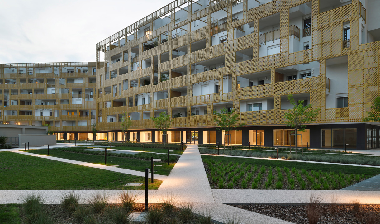 Modern residential complex at dusk with illuminated ground-floor units, landscaped courtyard, pathways, and dark bollard lights.