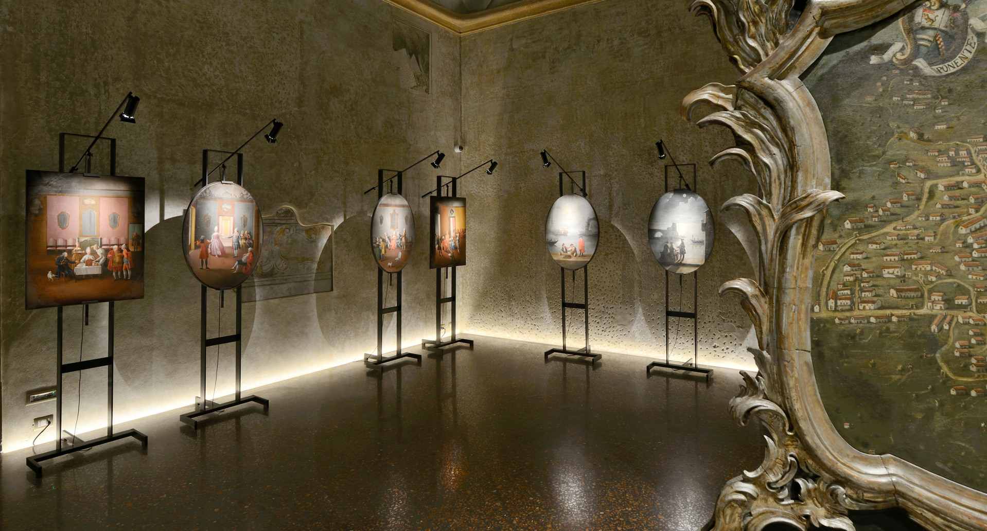 Sala espositiva con opere su cavalletti illuminate da faretti neri orientabili, striscia LED calda lungo la base delle pareti con texture.
