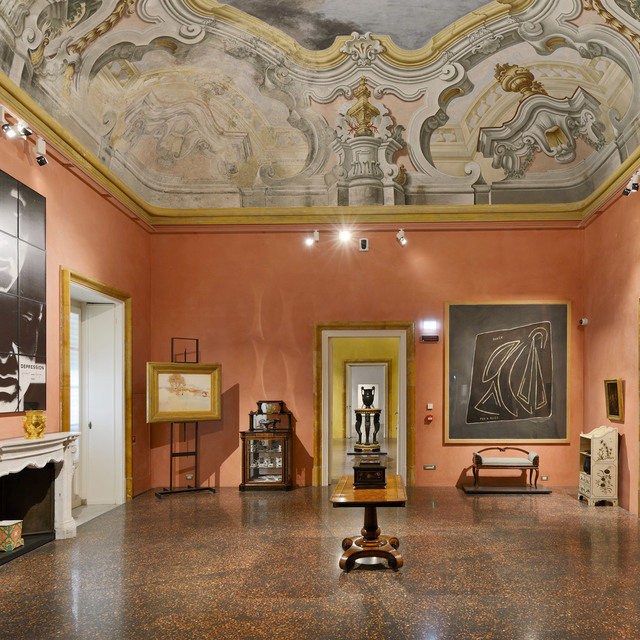 Sala con soffitto affrescato barocco, pareti color salmone, faretti su binario che illuminano opere d'arte, mobili antichi e pavimento in terrazzo lucido.