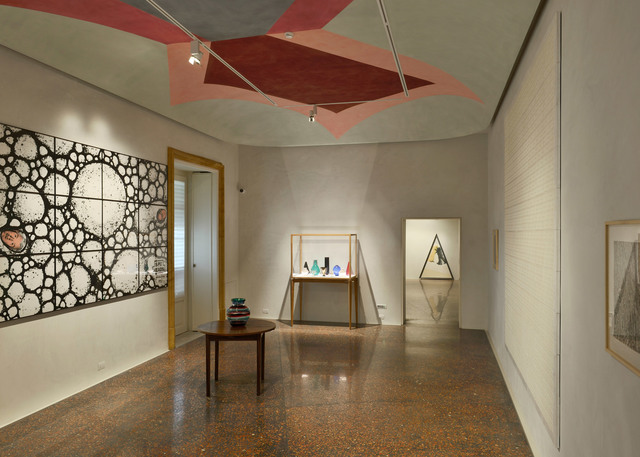 Interno di galleria con pavimento in terrazzo lucidato, opere d'arte alle pareti, vetrina con oggetti in vetro e faretti su binario su soffitto dipinto.