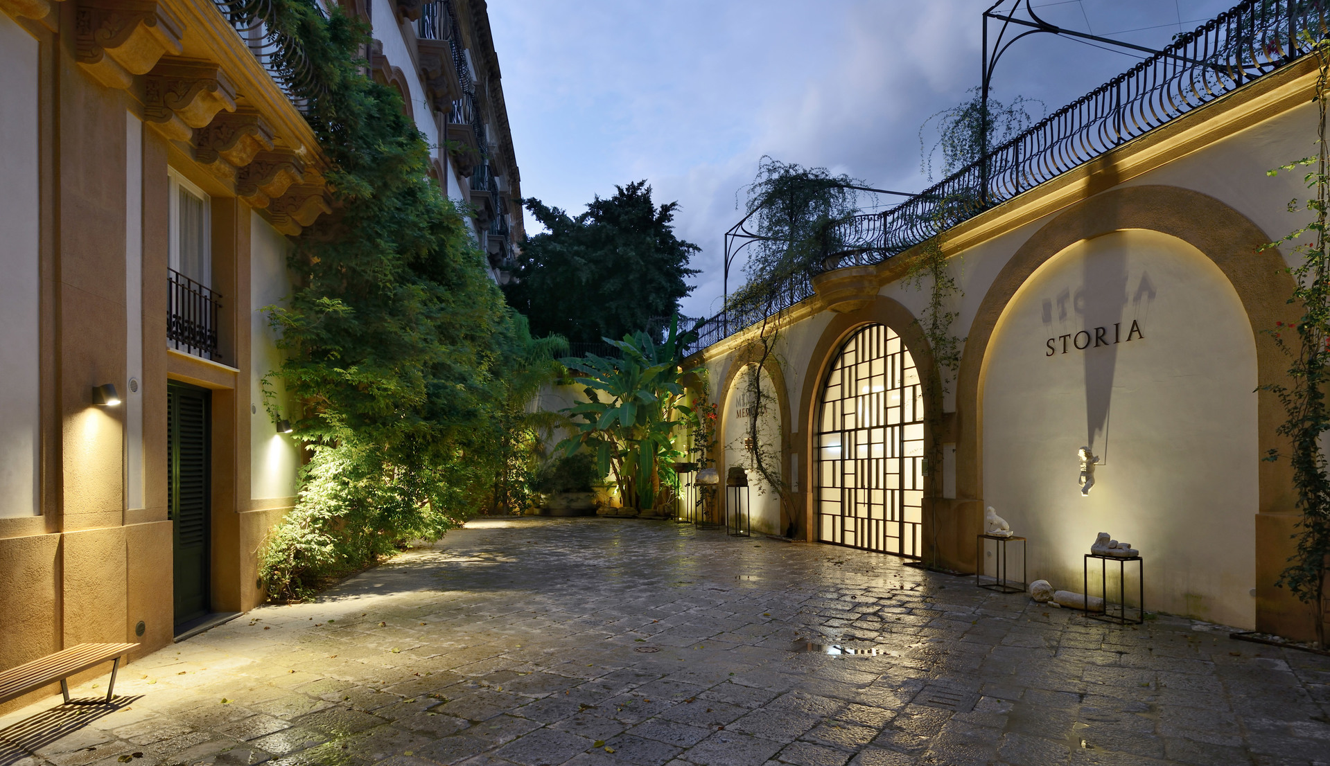 Elegante cortile al crepuscolo con facciata ad archi, rigogliosa vegetazione, pavimento in ciottoli e calde applique Artemide che illuminano lo spazio.