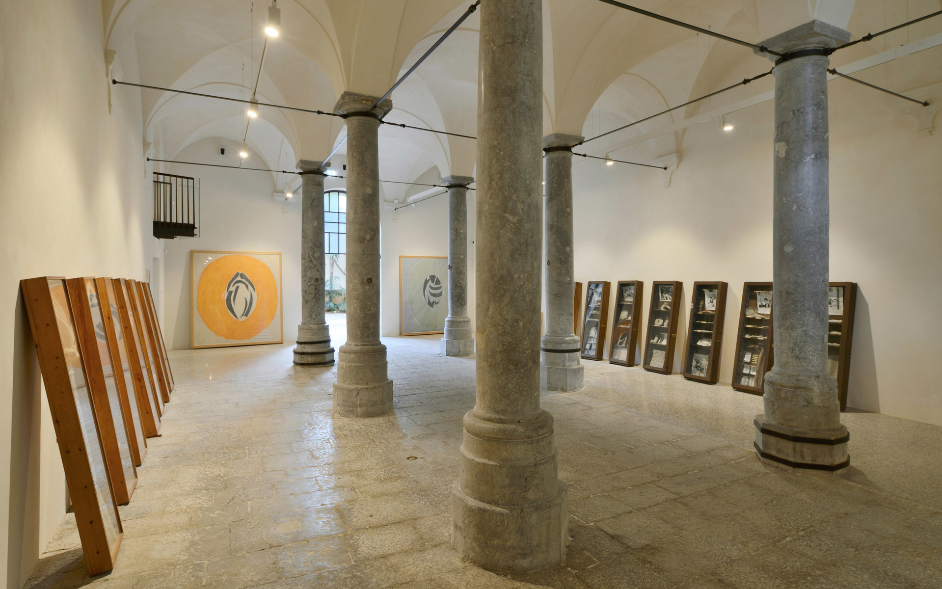 Spazio espositivo con soffitti a volta, colonne in pietra, cornici in legno appoggiate e opere d'arte illuminate da faretti a soffitto