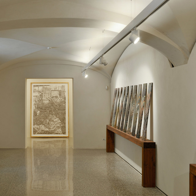 Galleria d'arte con soffitto a volta illuminato da faretti su binario Artemide, che valorizzano una grande opera e un'installazione scultorea su banco in legno