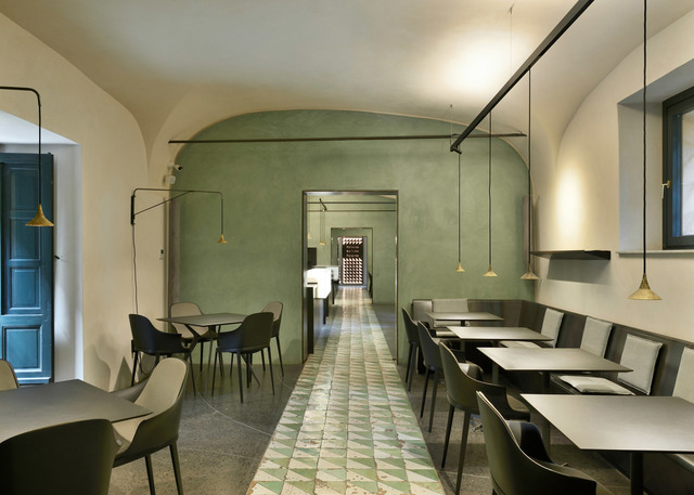 Interno di ristorante con soffitti ad arco, pareti verdi e lampade a sospensione coniche in ottone che illuminano tavoli e sedie scuri.
