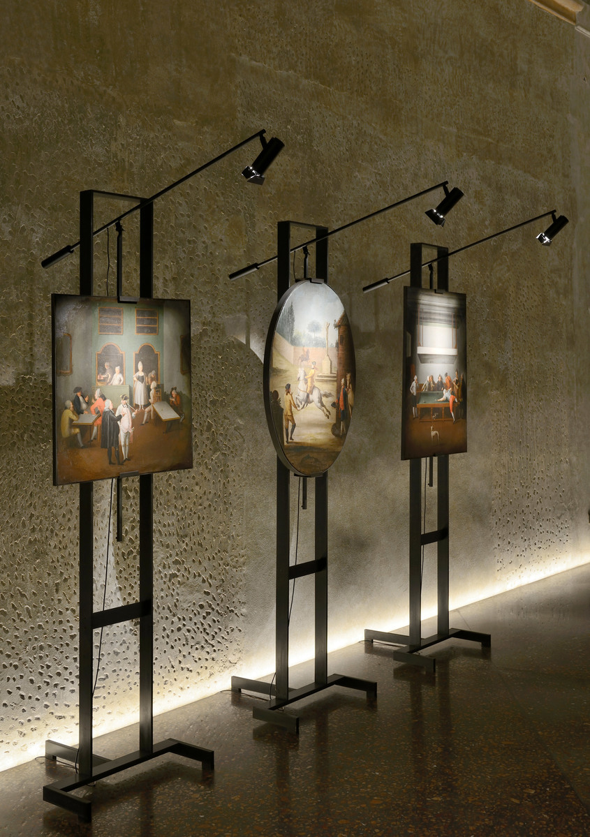 Tre supporti in metallo nero con opere d'arte illuminate in una galleria, ciascuno sormontato da un faretto Artemide orientabile che illumina i pannelli.
