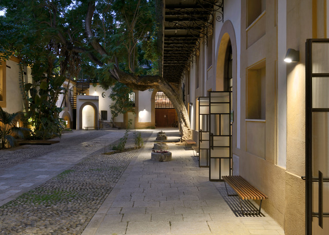Cortile storico al crepuscolo con lampade Artemide a parete che illuminano percorsi in pietra, facciate ad arco e scaffalature metalliche.