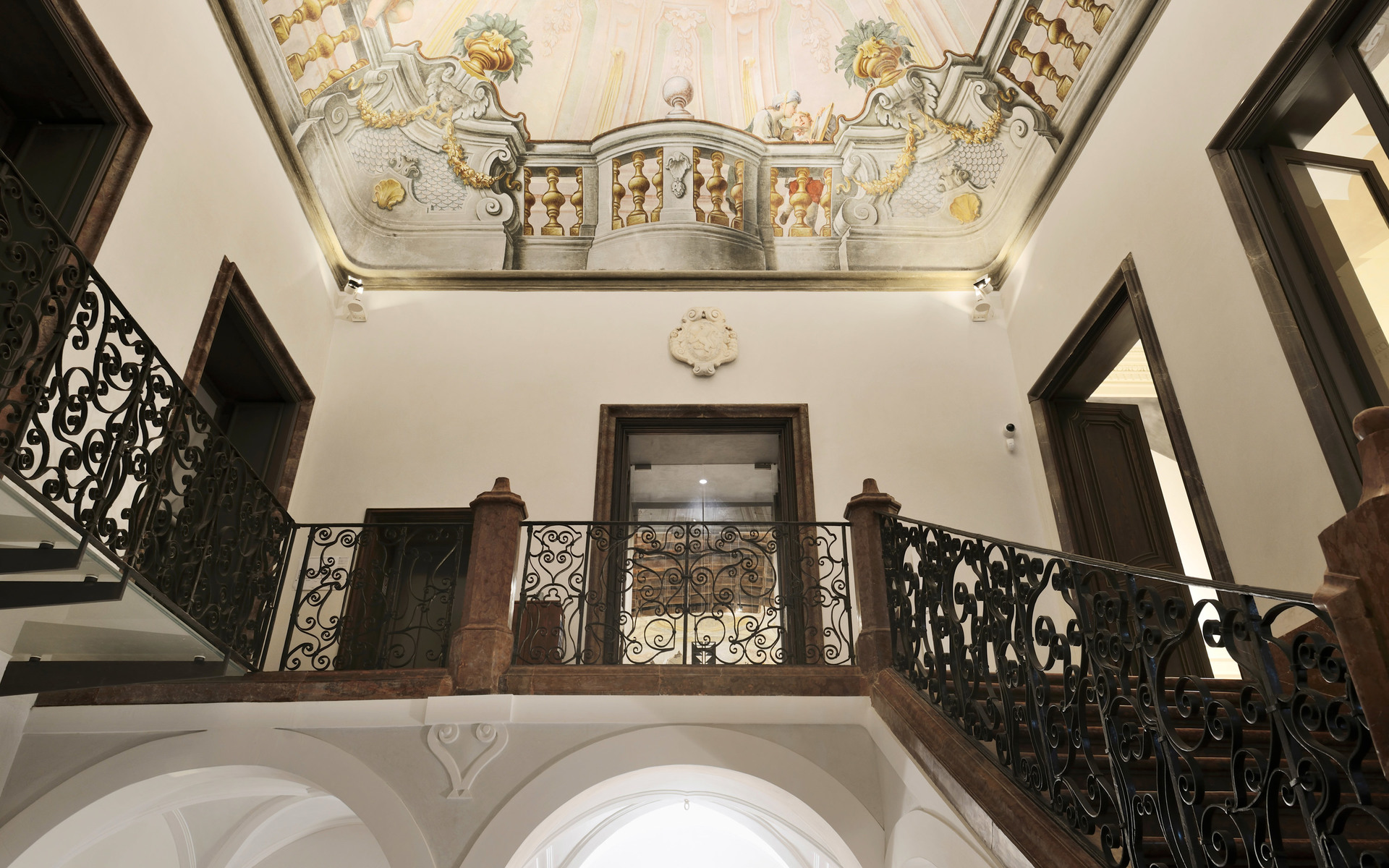 Interno di palazzo storico con soffitto affrescato ornato, scala con balaustra in ferro battuto e illuminazione d'ambiente calda su pareti bianche