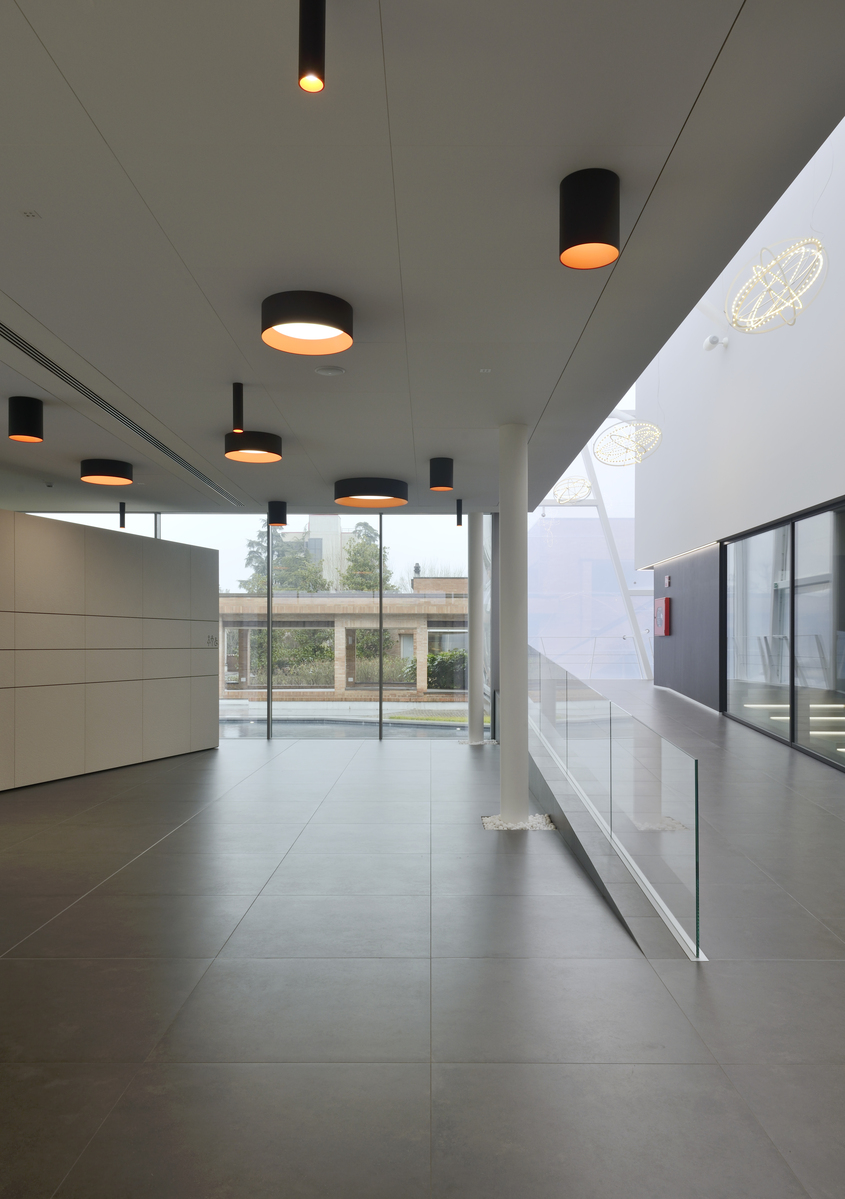 Corridoio moderno con multiple luci a soffitto cilindriche nere con interni dorati caldi che illuminano uno spazio aperto luminoso.