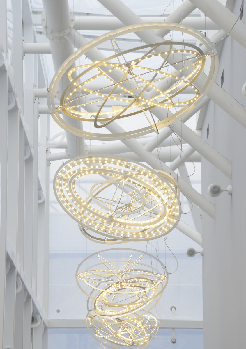 Tre grandi lampade a sospensione Artemide con anelli luminosi sovrapposti a LED bianco caldo, sospese in un atrio luminoso con colonne strutturali bianche