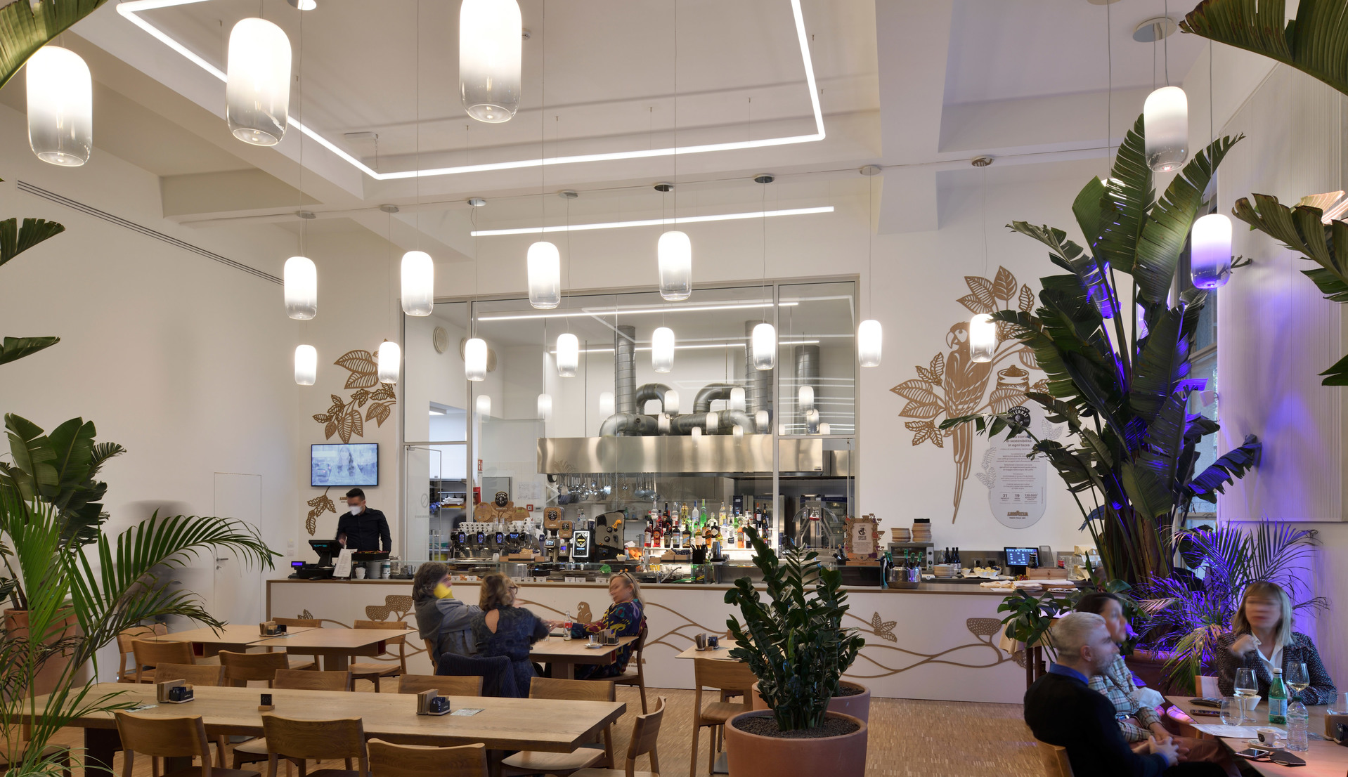 Interno di un moderno caffè con lampade a sospensione cilindriche bianche che illuminano la sala da pranzo, cucina aperta e piante tropicali.