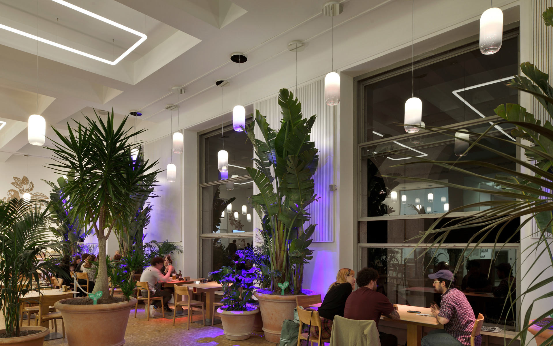 Interno di ristorante con lampade a sospensione cilindriche bianche Artemide, rigogliose piante tropicali e luci viola che creano un'atmosfera calda.