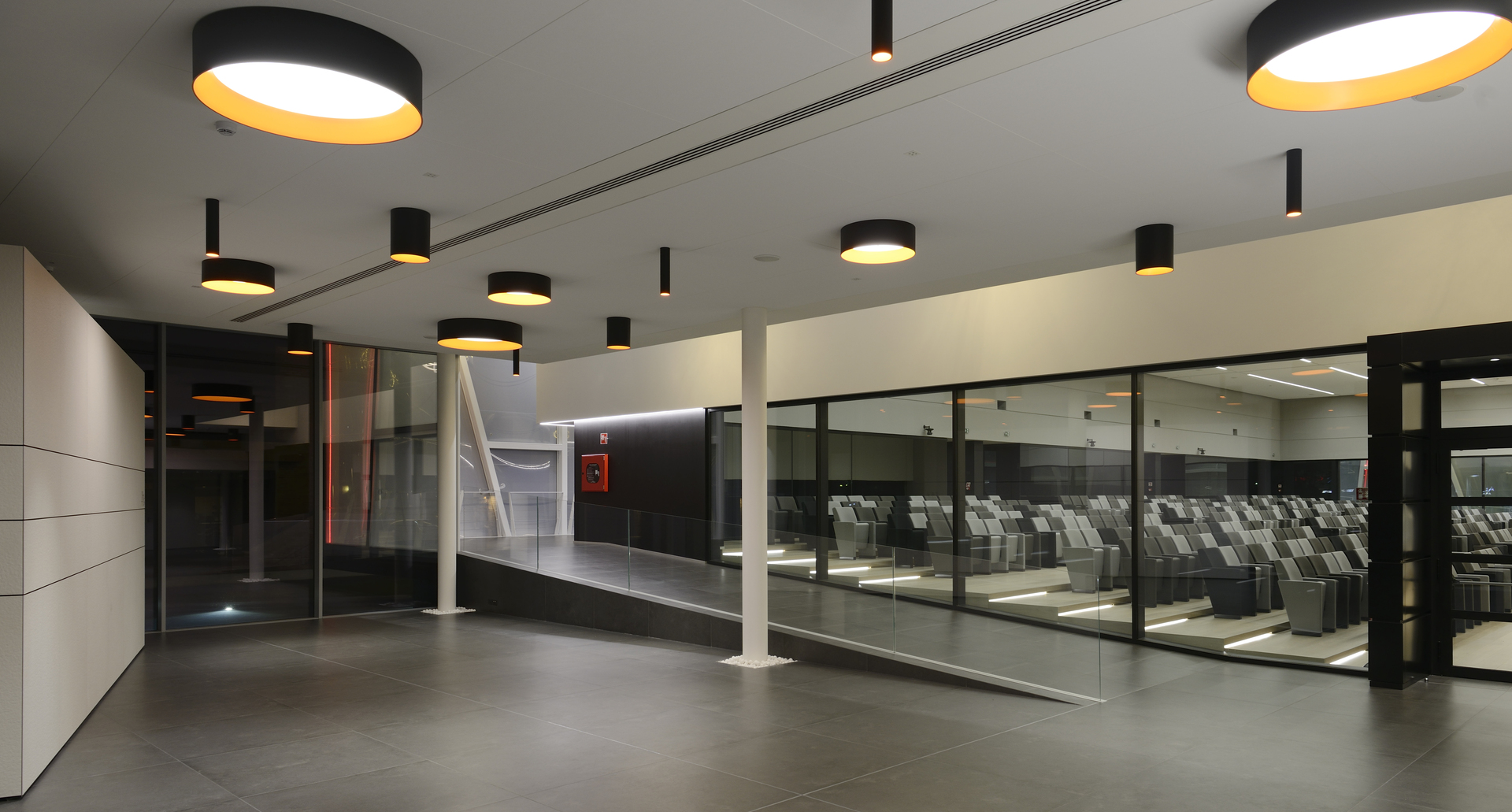 Ingresso moderno con lampade a sospensione cilindriche nere grandi e piccole con diffusori interni dorati, che illuminano uno spazio aperto con pareti in vetro.
