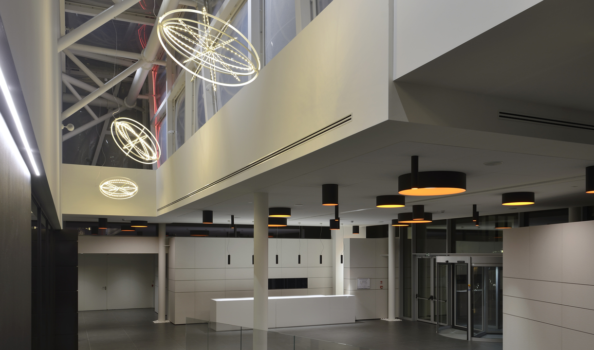 Ingresso moderno con lampade a sospensione ad anelli orbitali Artemide e plafoniere cilindriche nero-oro che illuminano un'ampia hall
