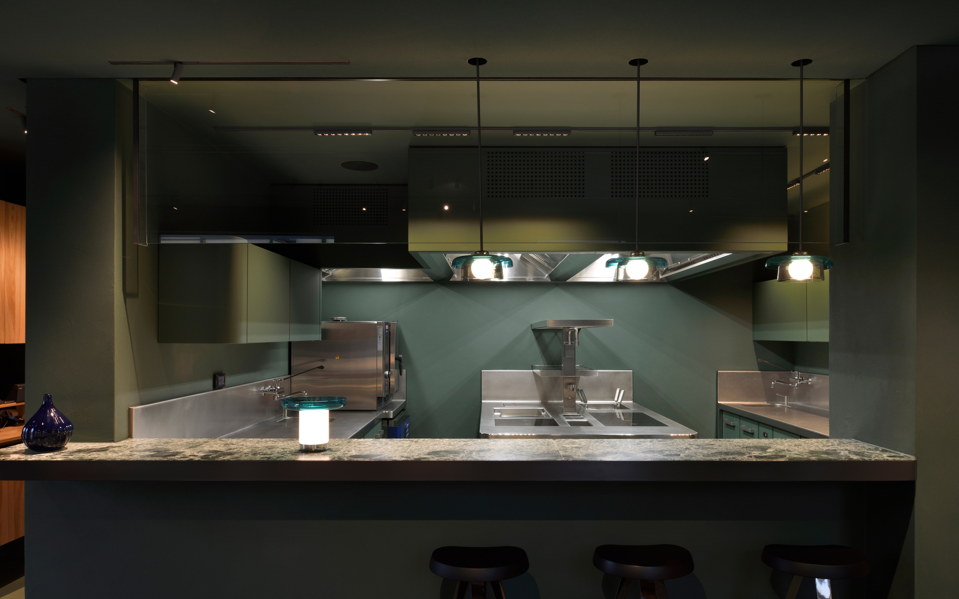 Modern professional kitchen with dark ambiance lit by three Artemide pendant lamps above a marble counter with bar stools.