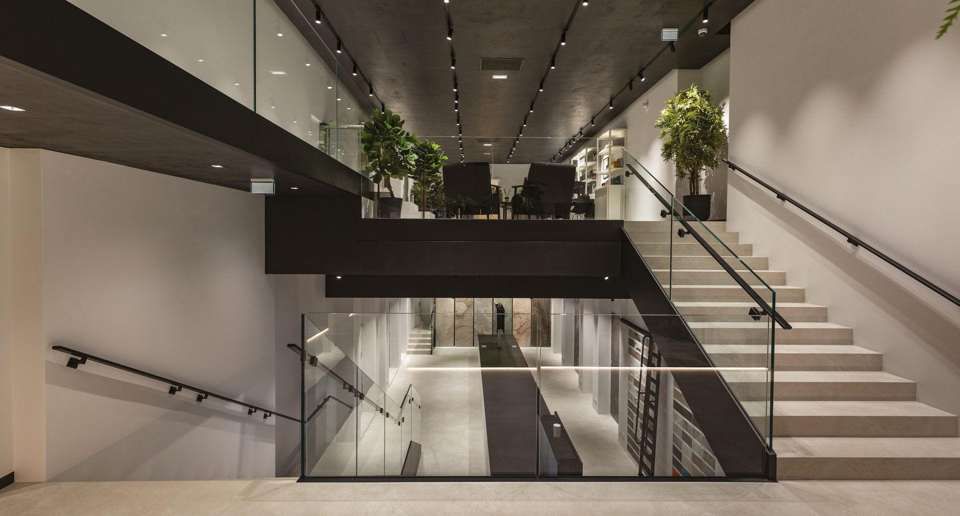 Modern multi-level interior with track lighting on dark ceiling, glass railings, white staircase, and green plants on upper floor.