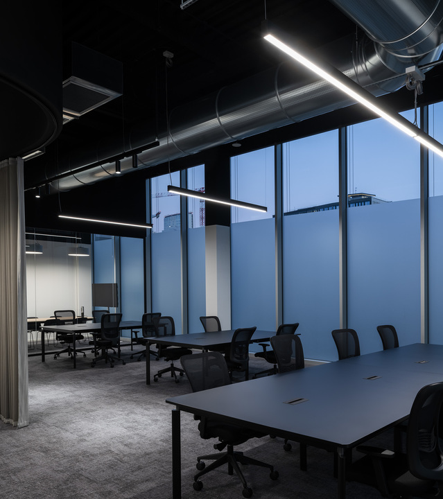 Ufficio open space moderno con lampade a sospensione LED lineari disposte diagonalmente su postazioni scure, finestre a tutta altezza al crepuscolo.