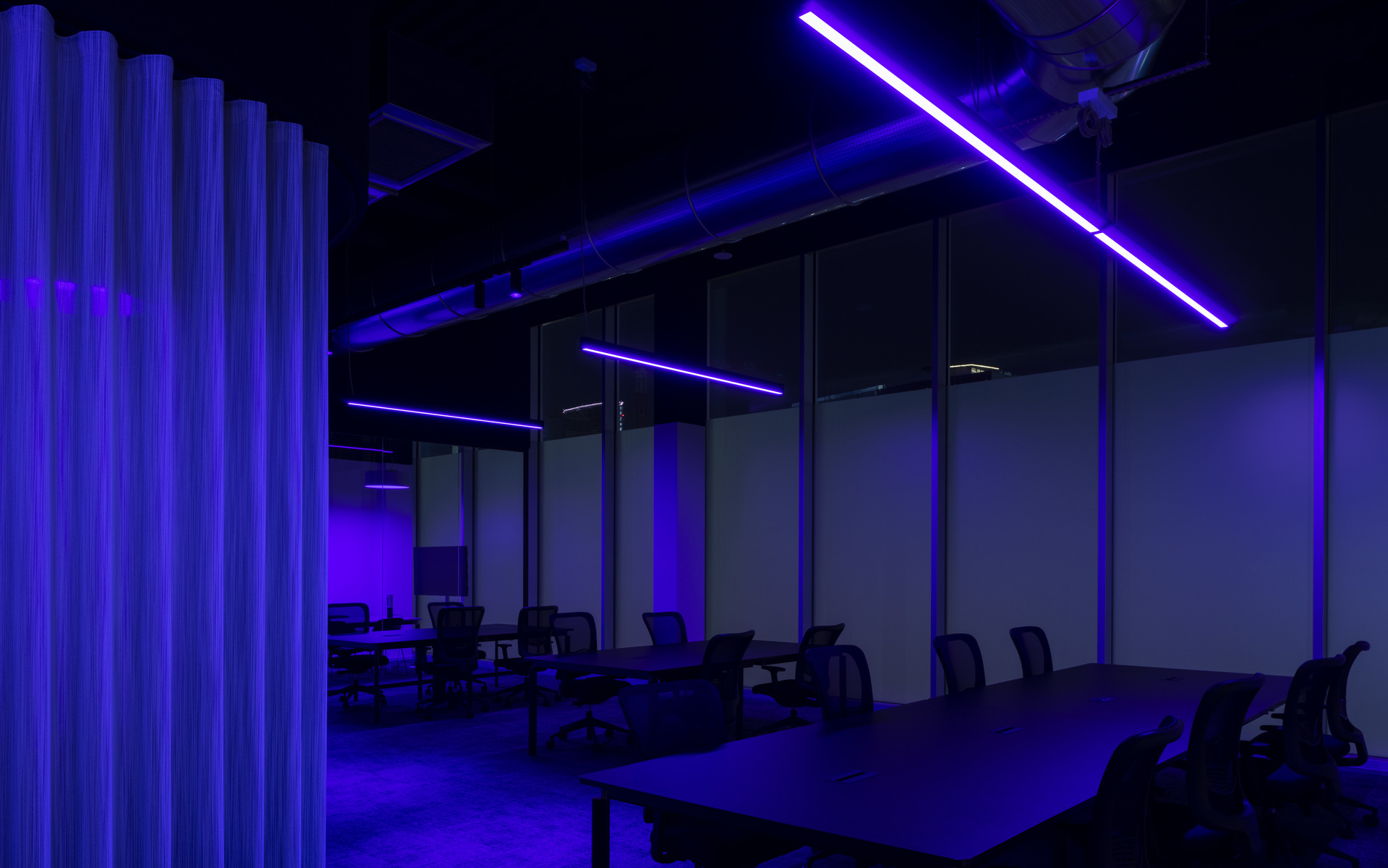 Ufficio scuro con luci LED lineari blu e viola a soffitto che formano una X, illuminando file di scrivanie e sedie