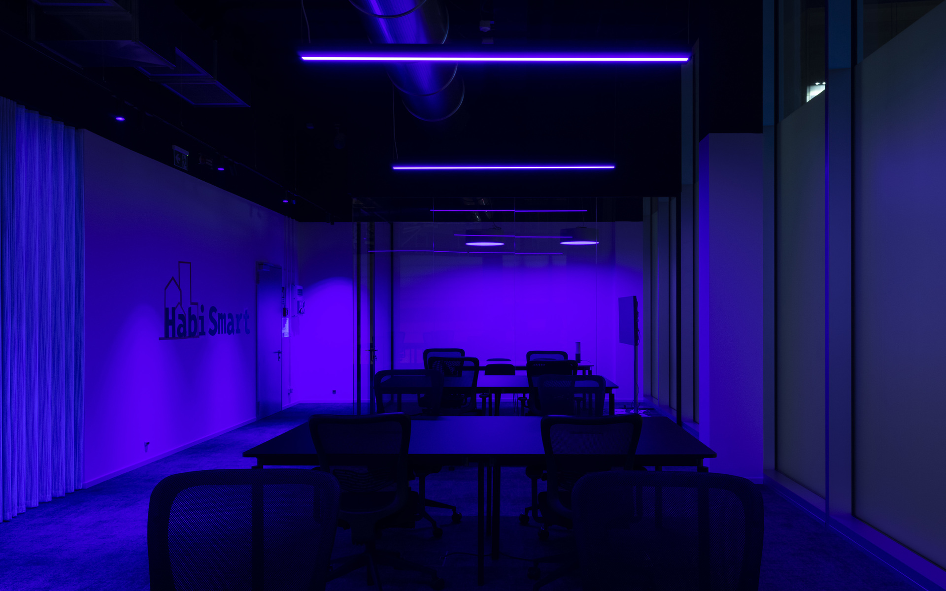Sala riunioni buia immersa in una vivida luce viola di lampade lineari LED a soffitto, con tavoli e sedie disposti in file.