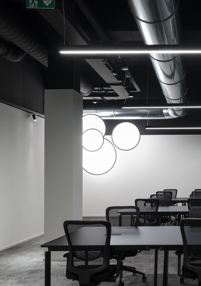 Spazio ufficio con lampade a sospensione circolari Artemide su parete bianca, strisce LED lineari a soffitto, tavoli scuri e sedie a rete.