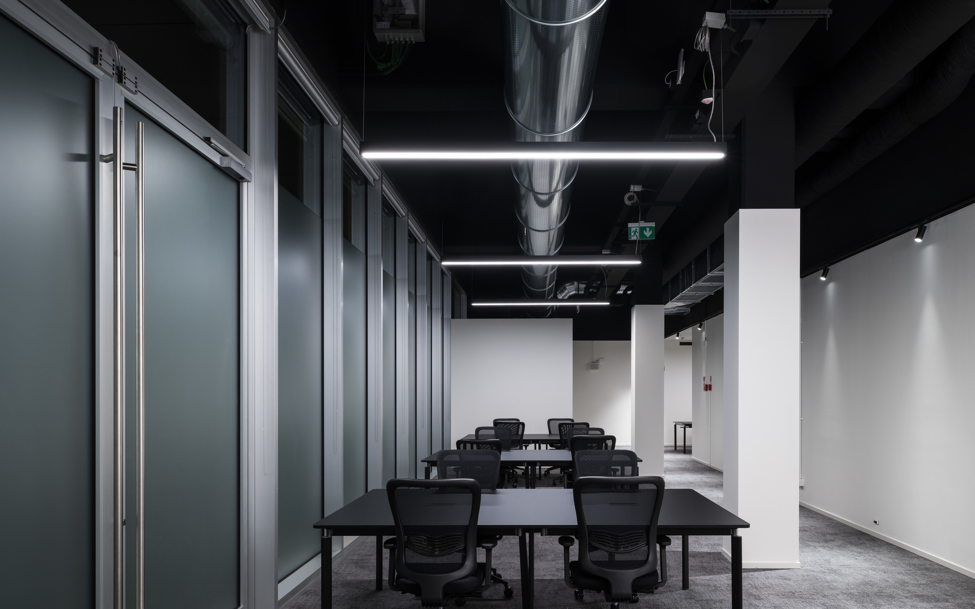 Sala conferenze moderna con soffitto nero, lampade a sospensione LED lineari che illuminano tavoli e sedie scuri, condotto a vista, parete divisoria in vetro.