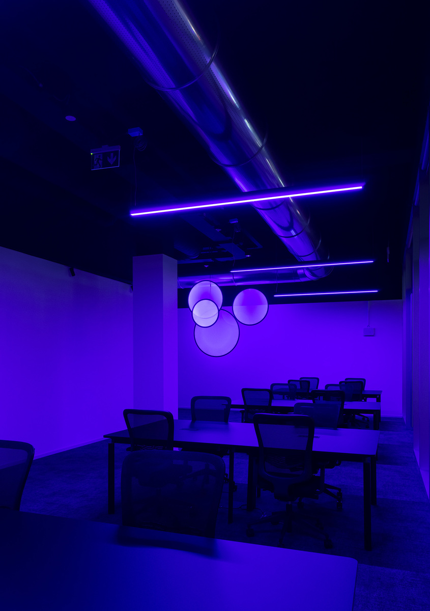 Spazio ufficio immerso in luce viola con lampade lineari a soffitto e tre lampade a sospensione sferiche luminose su parete scura.