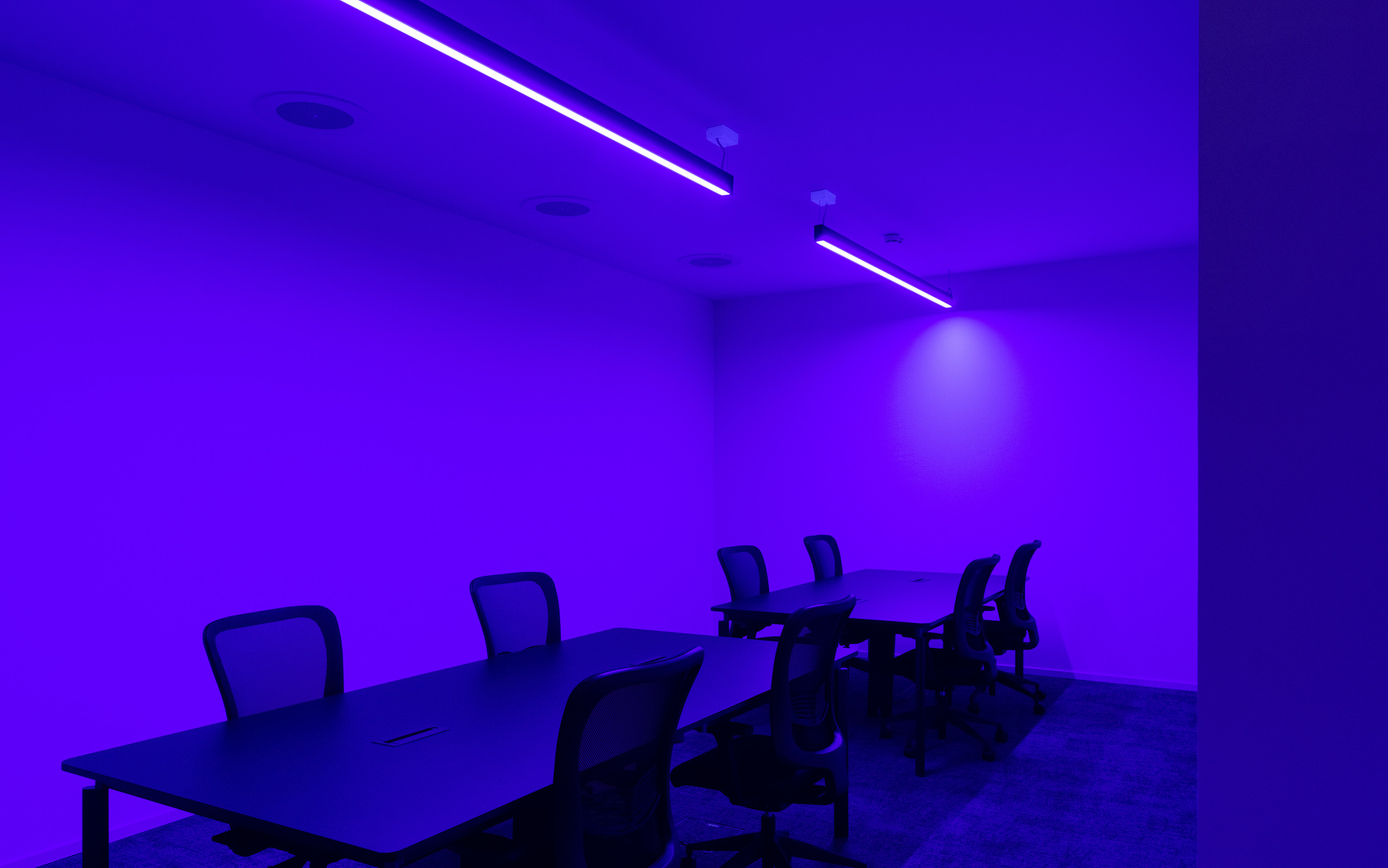 Sala conferenze immersa in luce viola da un apparecchio LED lineare a soffitto, con tavoli scuri e sedie a rete disposte per riunioni