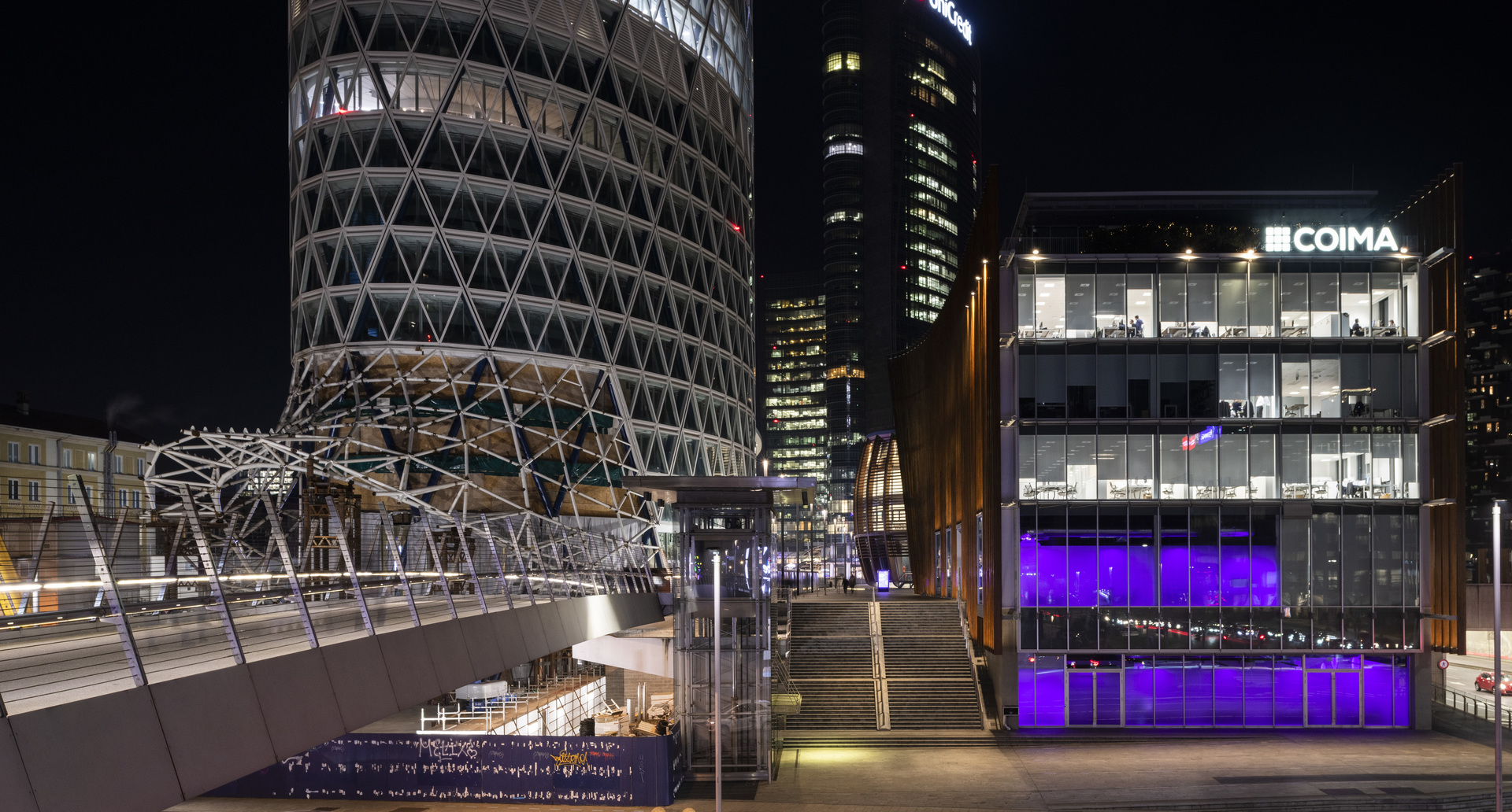 Paesaggio urbano di Milano di notte: edificio COIMA illuminato con luce viola, torre cilindrica con facciata geometrica e passerella sopraelevata