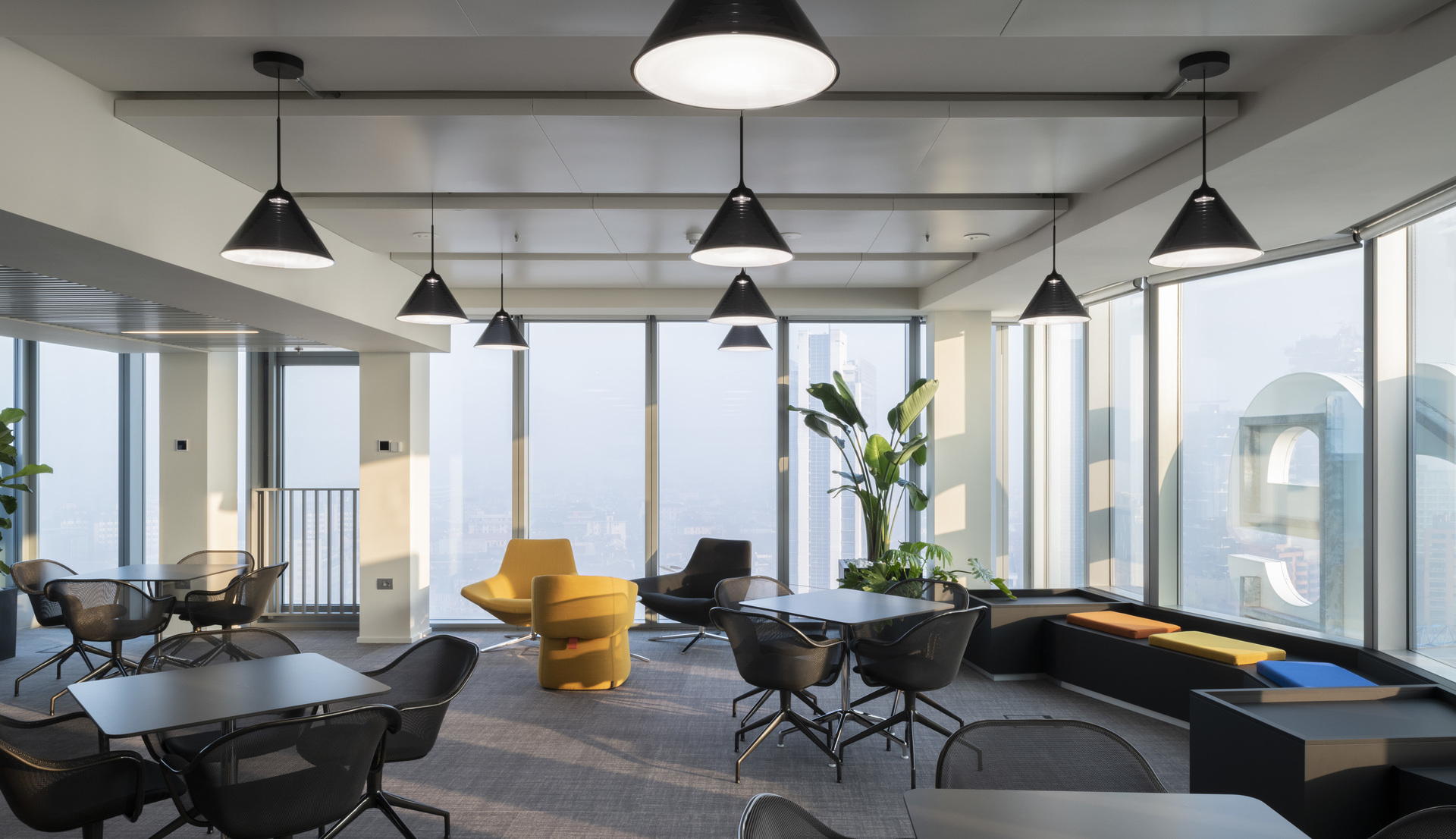 Modern office lounge with black conical pendant lights above dark tables, yellow accent chairs, and floor-to-ceiling windows with city views.