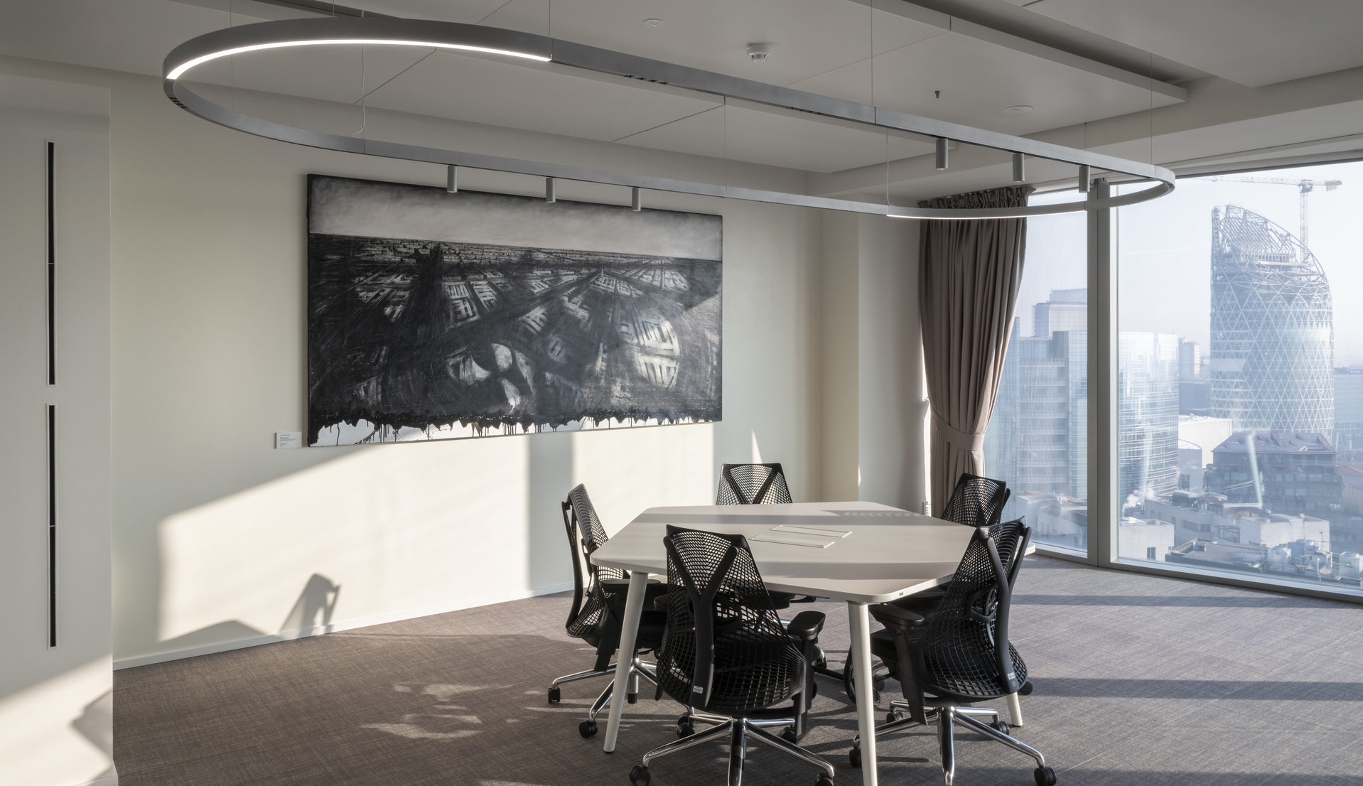 Modern conference room with oval Artemide pendant light ring above a round table, floor-to-ceiling windows overlooking a city skyline.