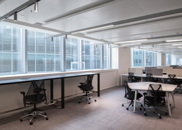 Modern open-plan office with Artemide ceiling track lights, long workbenches by windows, and clustered white desks with ergonomic chairs