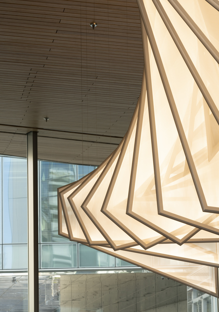Large sculptural pendant lamp with layered geometric panels in warm white, creating a spiral cone shape, suspended in a modern interior.