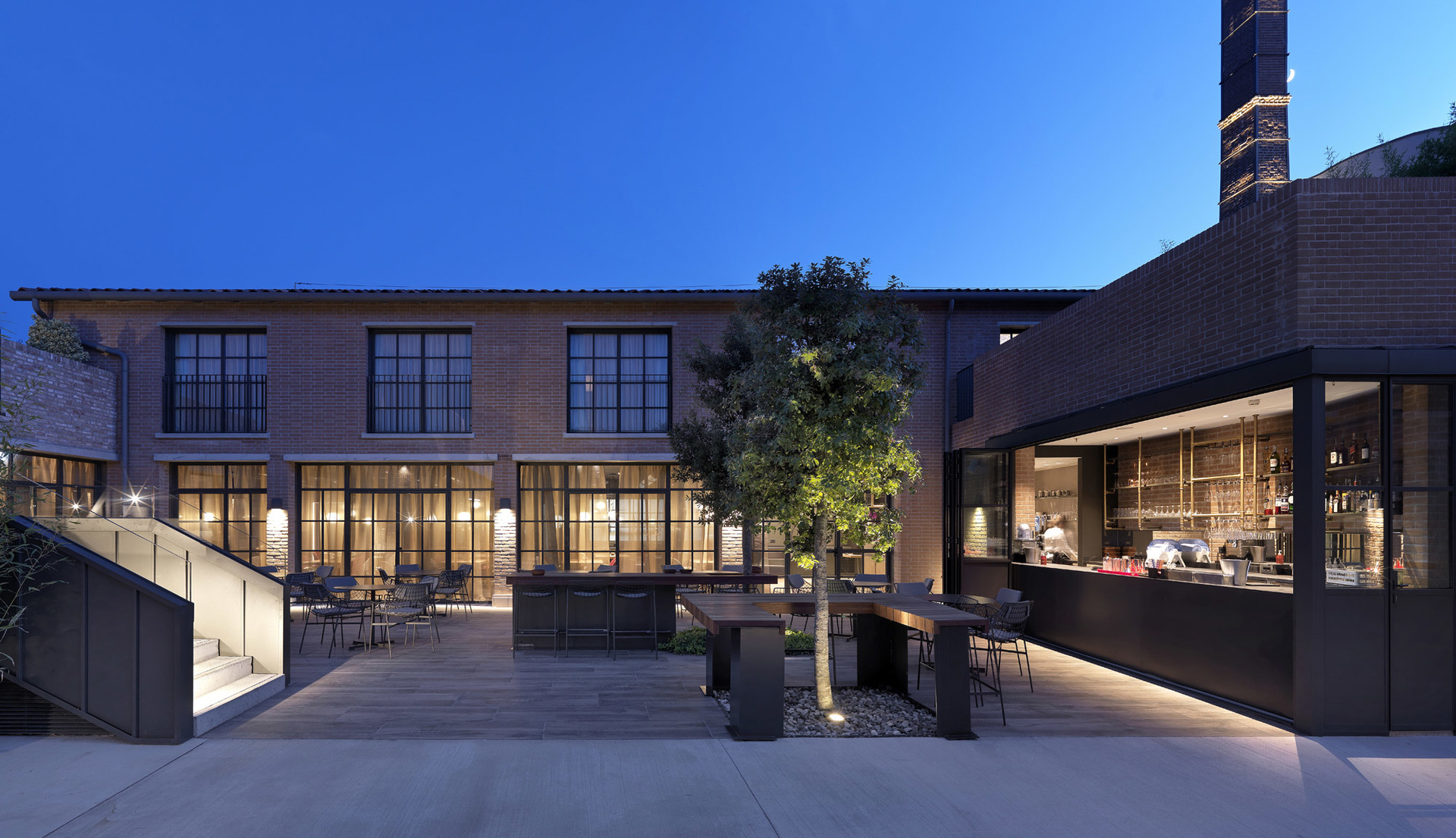 Cortile di edificio industriale in mattoni al crepuscolo con illuminazione esterna calda che illumina alberi, area pranzo e terrazza bar.