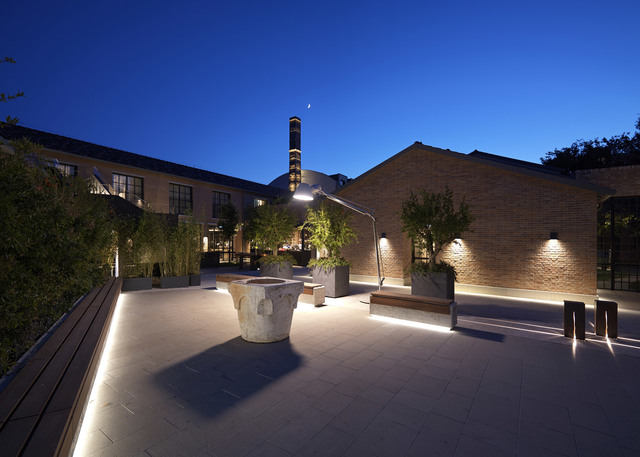 Cortile industriale in mattoni al crepuscolo con illuminazione LED a terra, applique a parete, paletti luminosi e grande lampada da terra articolata tra gli alberi.