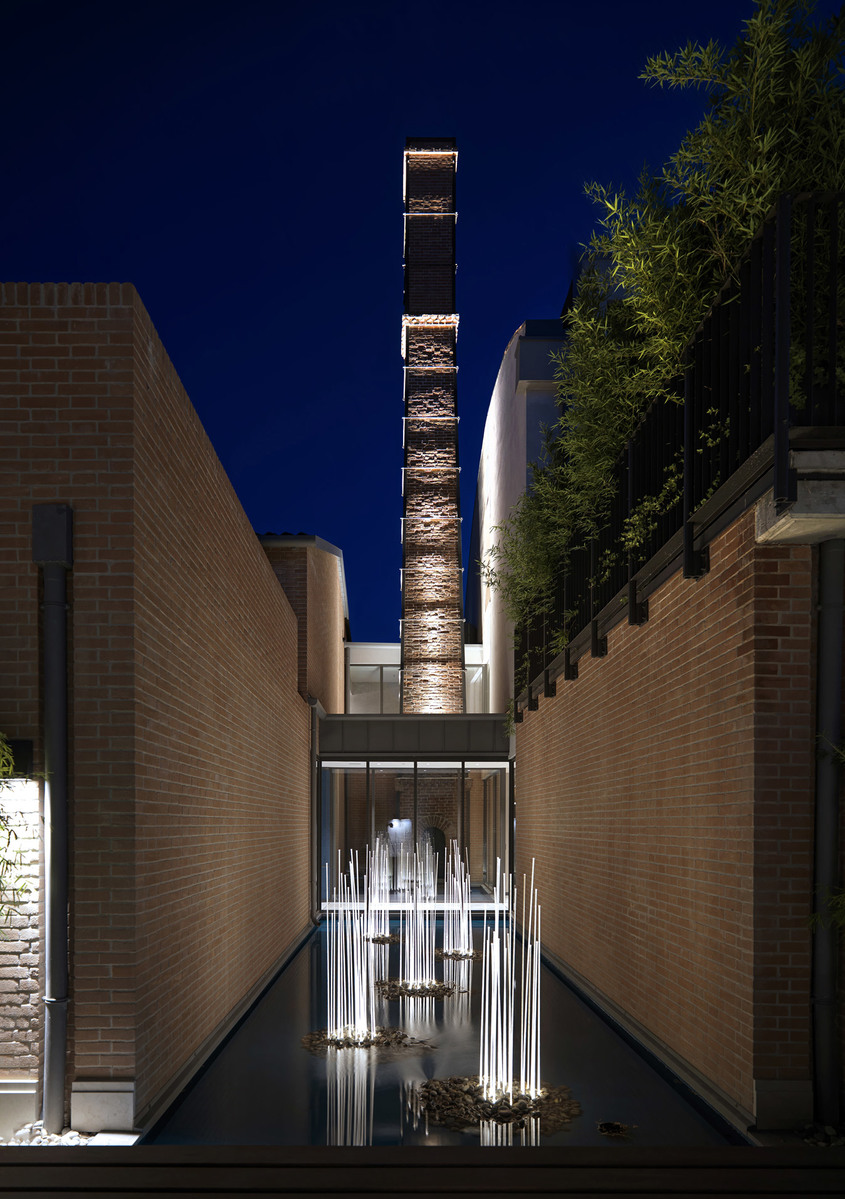 Cortile notturno con torre-camino in mattoni illuminata, vasca riflettente e installazioni luminose verticali tra muri in mattoni