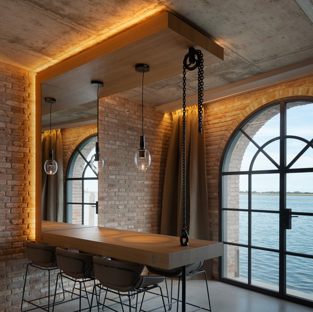 Area pranzo industriale con lampade a sospensione a globo in vetro sopra un tavolo bar in legno, illuminazione LED calda a cornice, pareti in mattoni e finestre ad arco con vista sull'acqua.