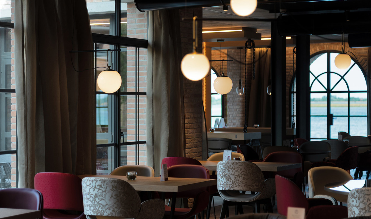Interno di ristorante con lampade a sospensione sferiche in vetro bianco sopra tavoli in legno e sedie colorate, grandi finestre con vista sul lungofiume
