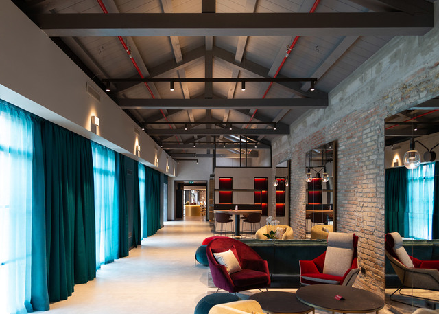 Lounge di hotel moderno con soffitto a volta, illuminazione su binario, tende color teal, muri in mattoni a vista e aree salotto colorate.
